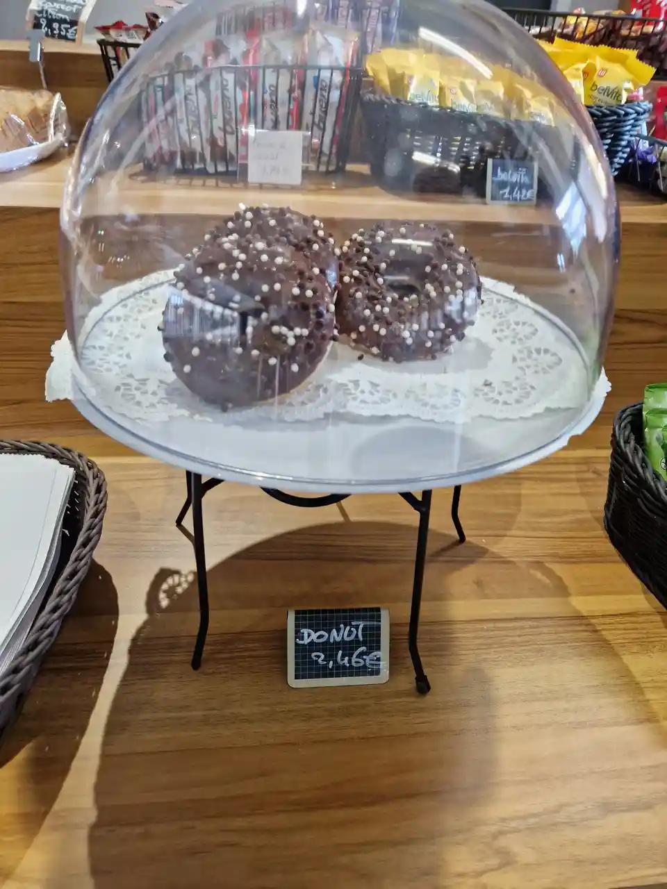 Two chocolate-glazed donuts with white and dark sprinkles sit under a clear glass dome on a white doily, displayed on a…