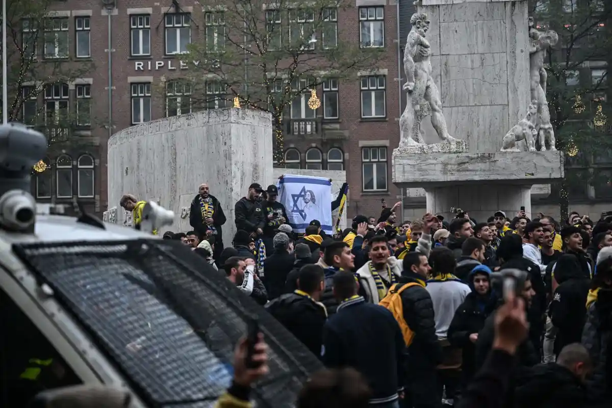 A crowd of people, many wearing yellow and black scarves, gathers around a large stone monument with statues in a city…
