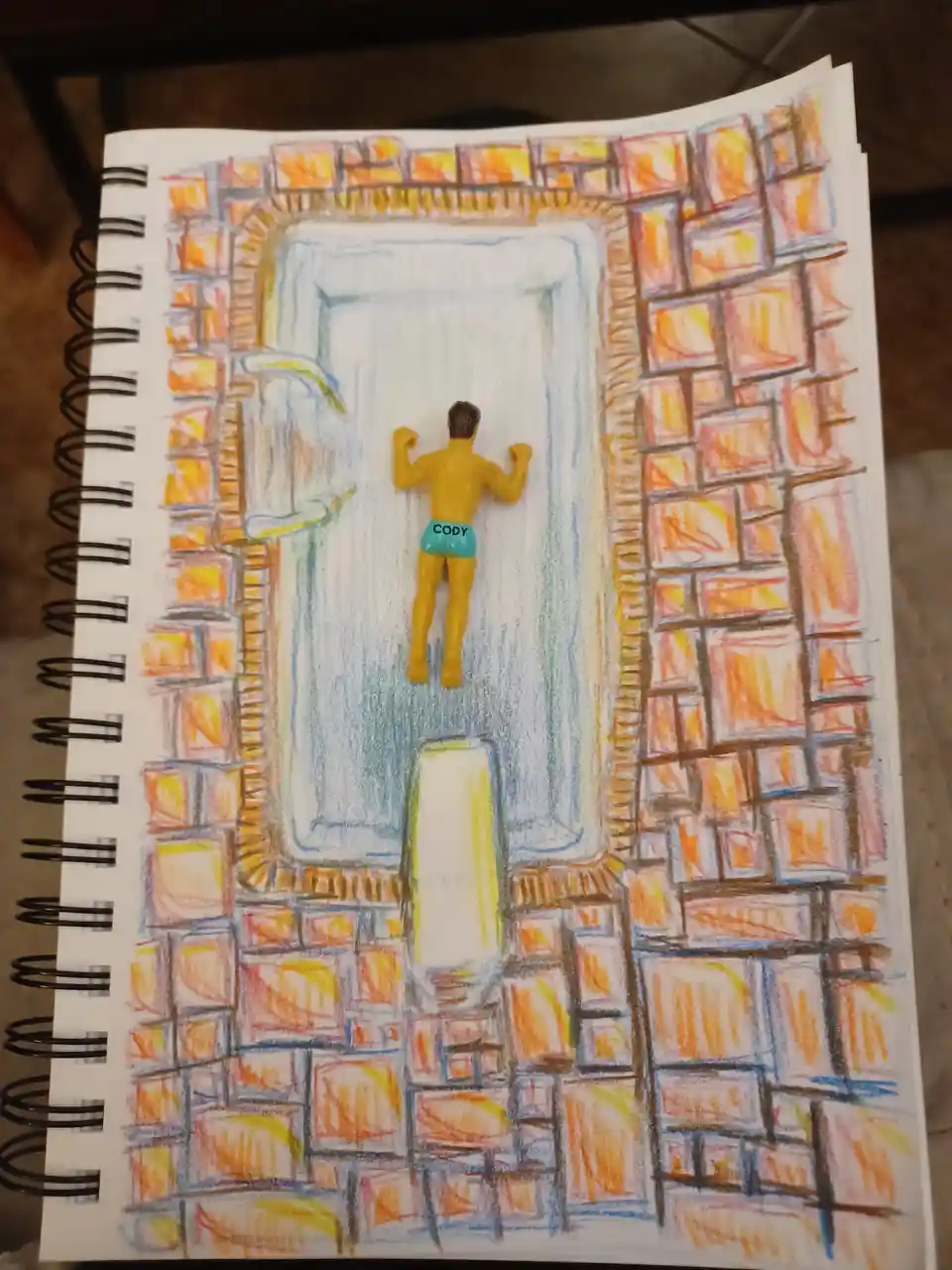 A hand-drawn illustration in a spiral-bound notebook depicts a small yellow plastic figurine of a man wearing blue swim…