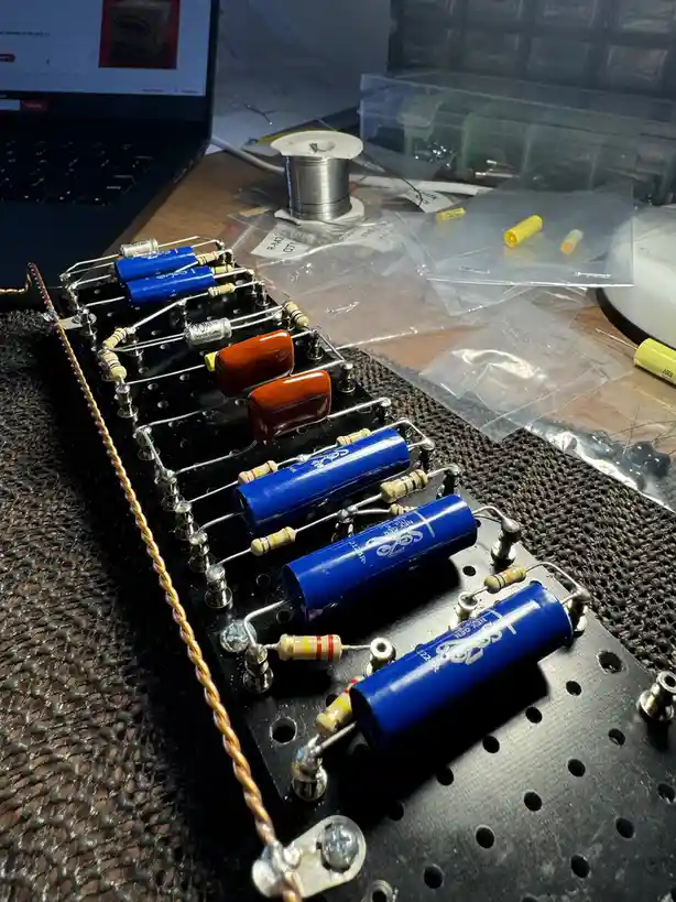 A close-up photo shows a black perforated soldering board with several blue cylindrical capacitors and smaller brown/orange…