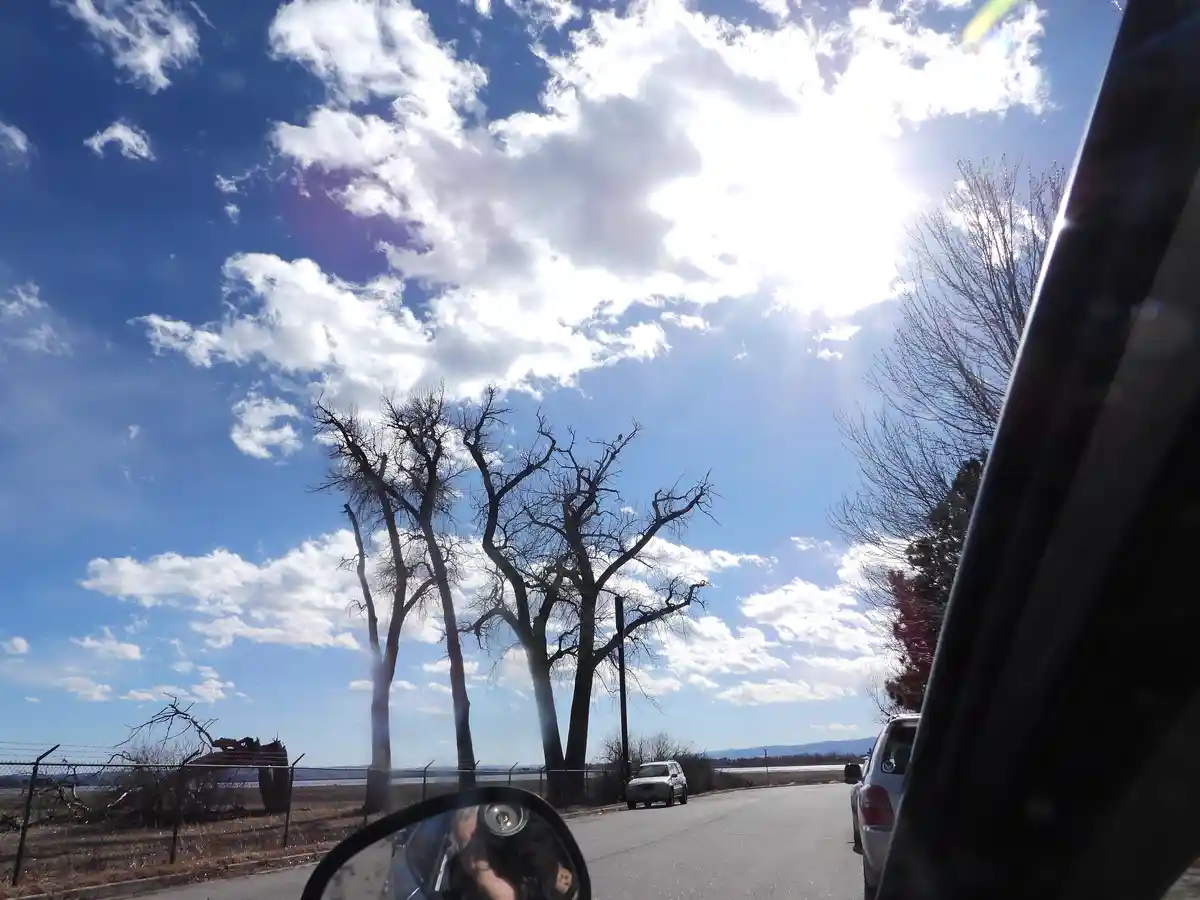 A photo taken from inside a car shows a bright blue sky with scattered white clouds and the sun shining brightly. Bare…