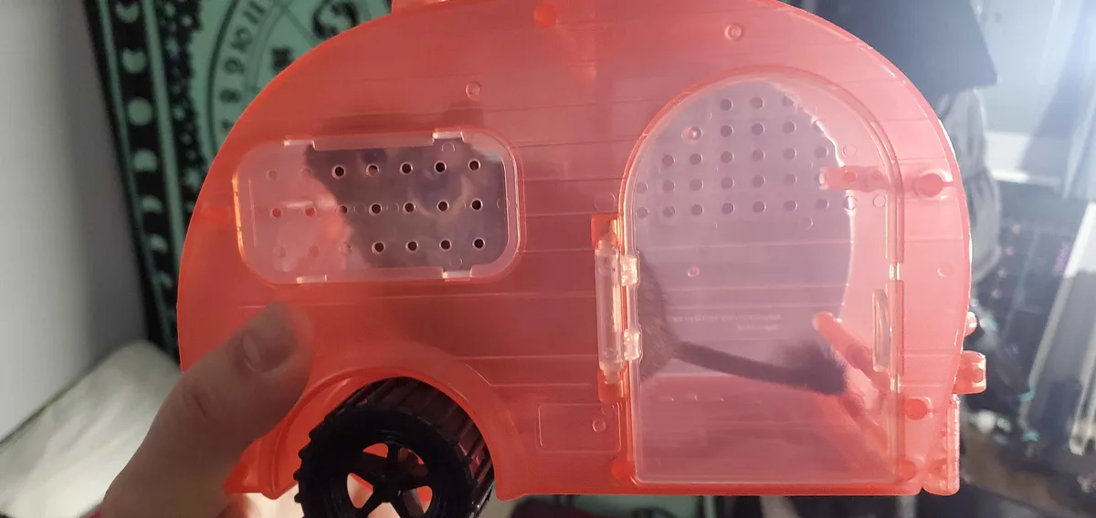 A hand holds a translucent pink plastic toy camper with a black wheel, its door open to reveal a mouse inside. The mouse is…