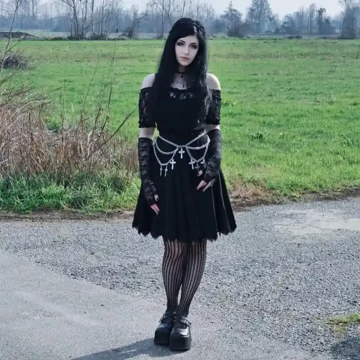 A woman with long black hair and pale makeup stands in a grassy field wearing a black goth outfit with lace, a cross-adorned…