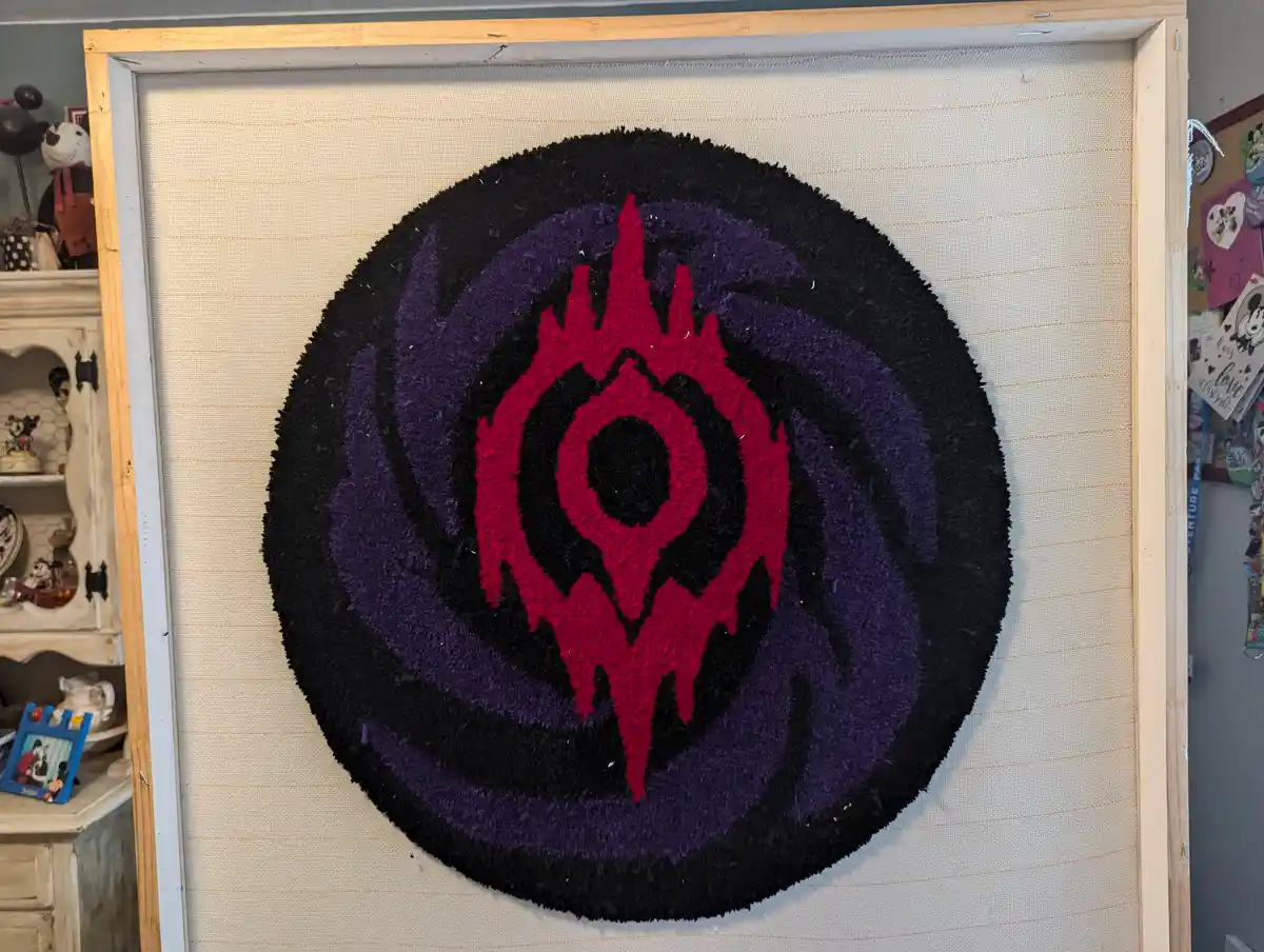 A handmade rug-hooked artwork of the World of Warcraft Legion logo, featuring a red and purple eye-like symbol on a black…
