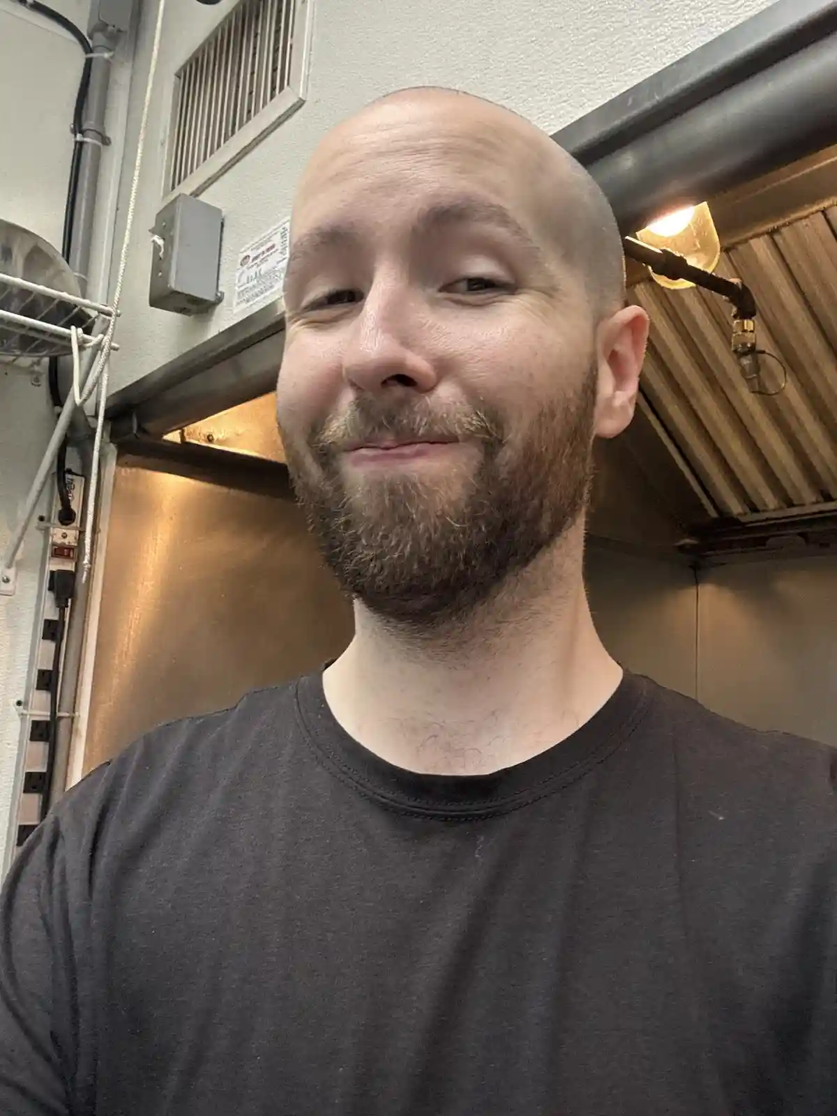 A bald man with a beard wearing a black t-shirt smiles smugly at the camera in what appears to be a commercial kitchen, with…