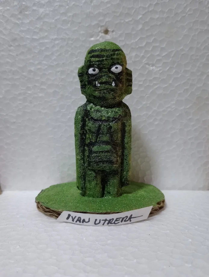 A handmade green figurine resembling a zombie or monster with wide eyes and fangs, standing on a green base with a…
