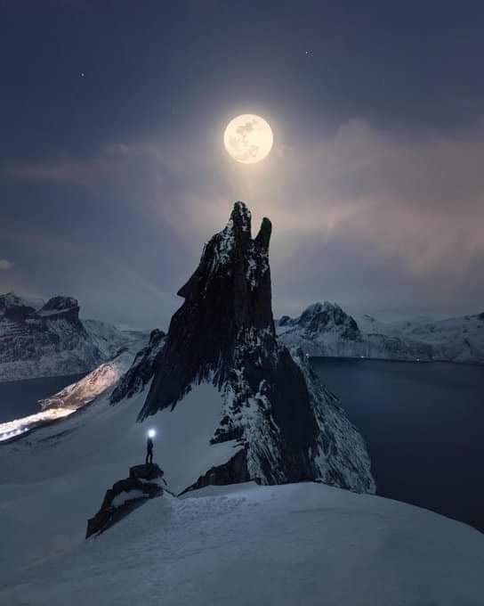 A lone person stands on a snowy mountain peak at night, holding a flashlight that illuminates the immediate area. A large…