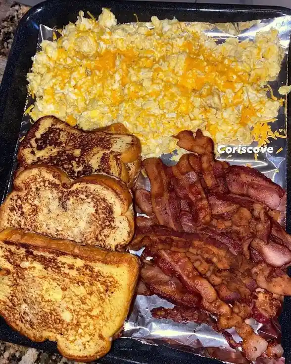 A black baking tray lined with foil holds a large breakfast spread: scrambled eggs topped with shredded cheddar cheese…