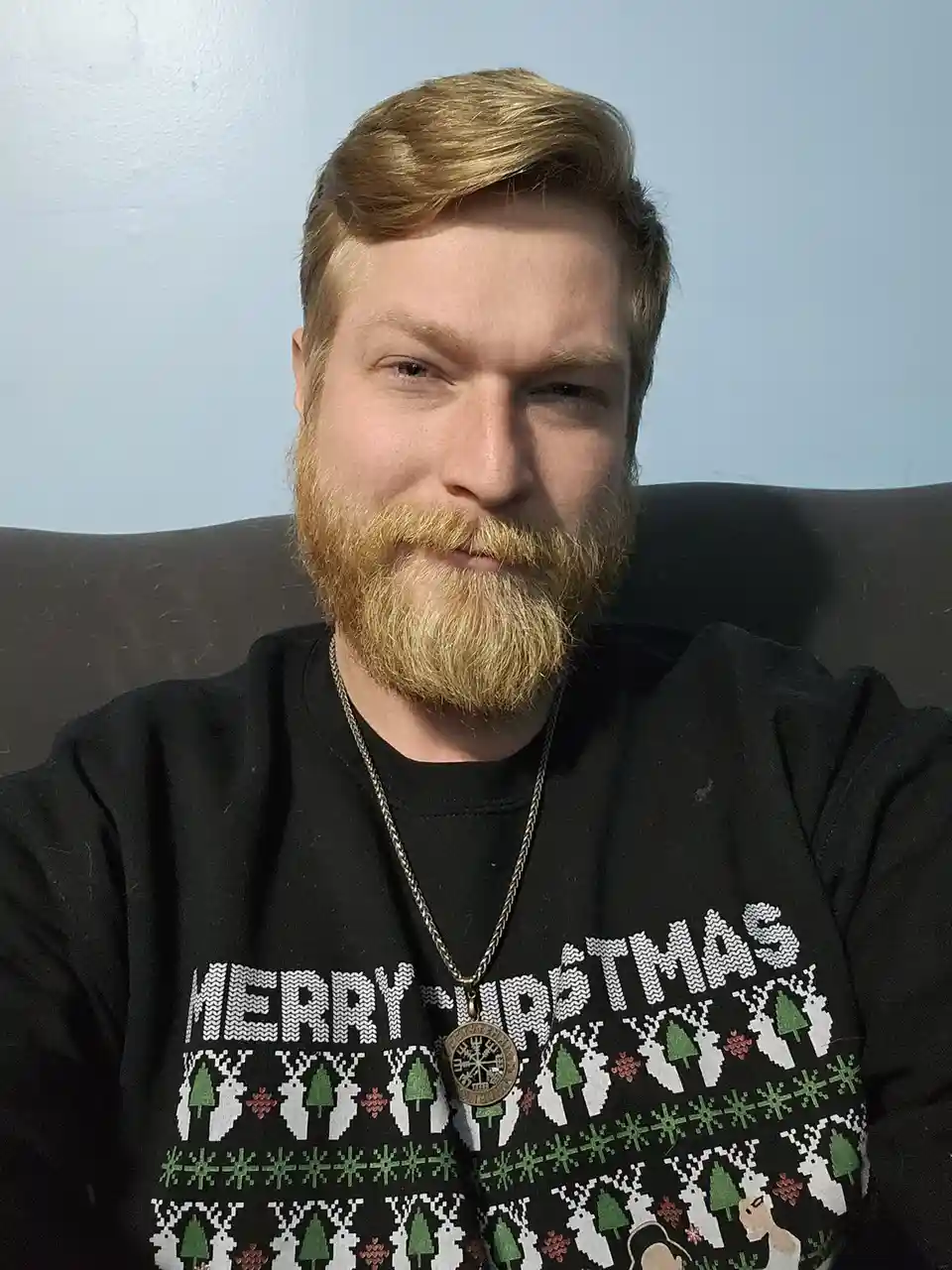 A man with a reddish beard and hair sits on a dark couch wearing a black Christmas sweater with white, green, and red…