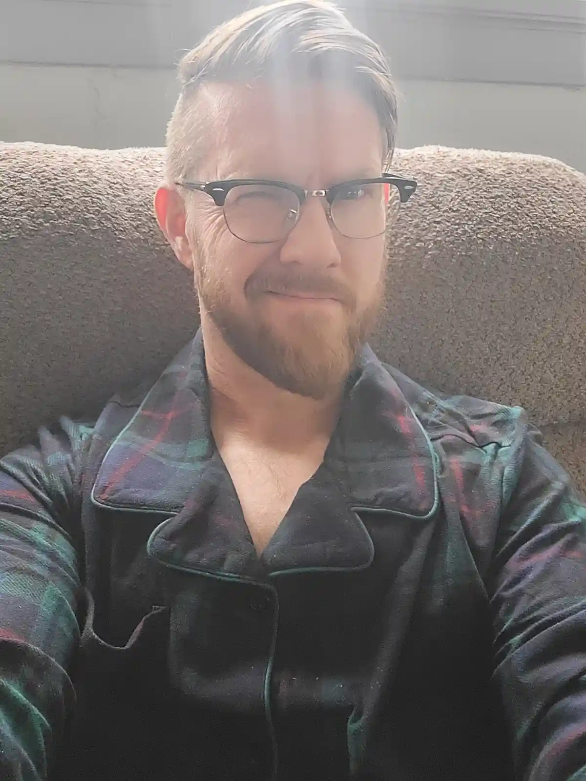 A man with glasses and a beard, wearing plaid pajamas, sits on a couch with a smug, self-satisfied expression. Bright…