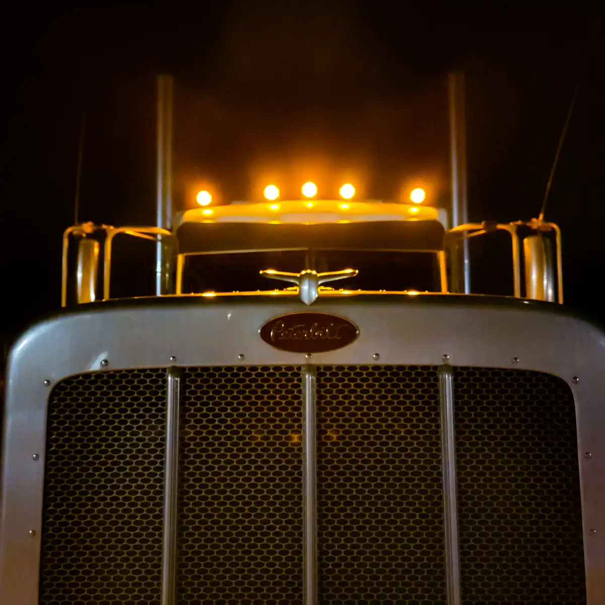 A close-up nighttime photo of the front of a Peterbilt semi-truck, showing its illuminated grille, chrome accents, and…