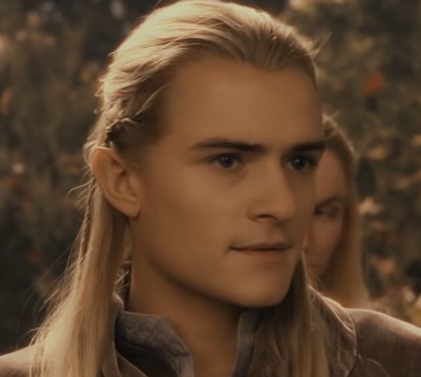 A close-up of a man with long blonde hair tied back, looking slightly to the side with a neutral expression. Behind him…