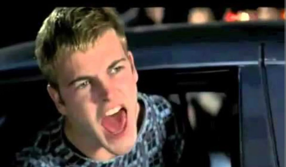 A young man with short blond hair, wearing a patterned shirt, is shown inside a car at night, screaming with his mouth wide…