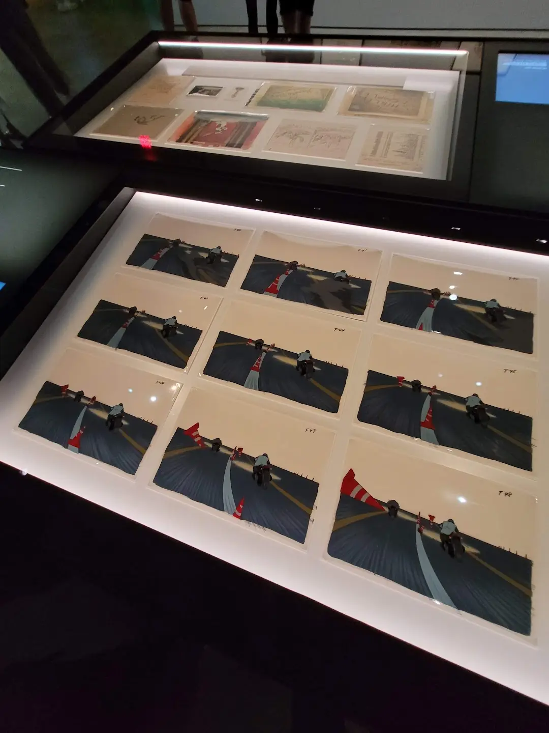 A museum exhibit displays a grid of hand-drawn animation cells showing a motorcycle rider leaning into a turn, with frames…