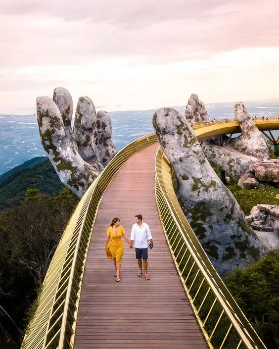 A couple walks hand-in-hand along a golden, elevated wooden bridge that arches between two giant, moss-covered stone hands…