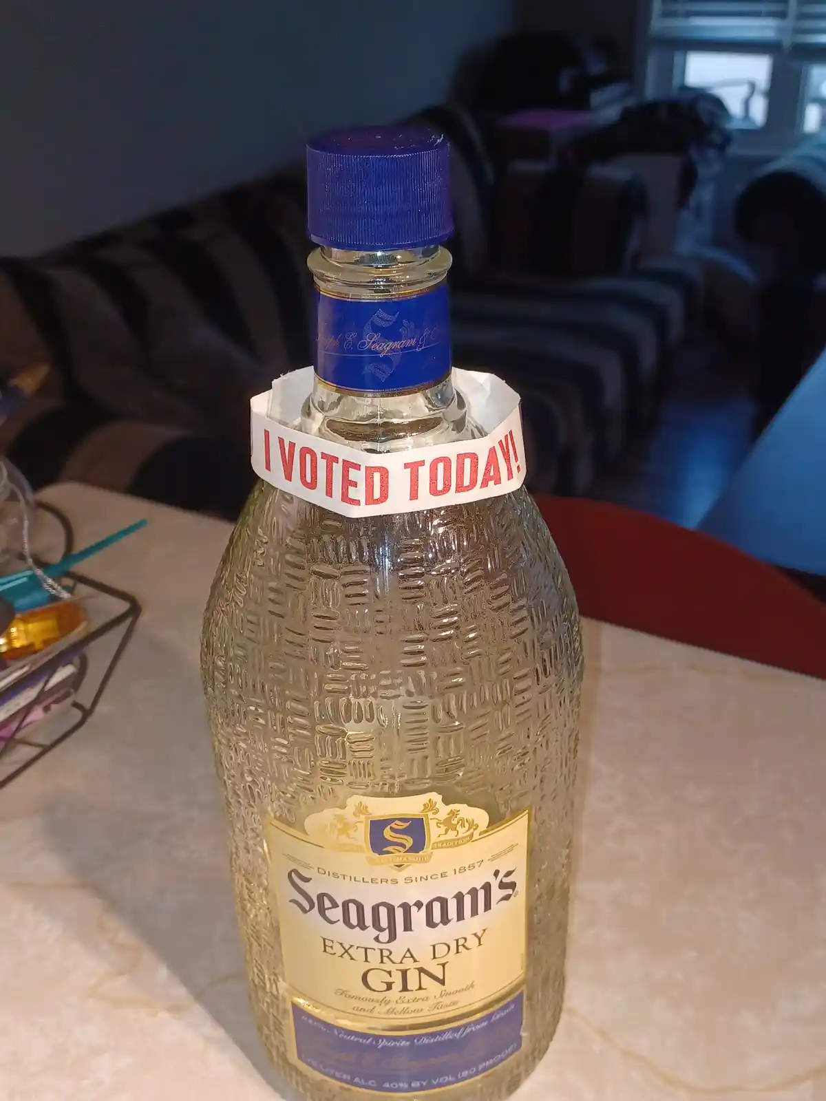 A bottle of Seagram's Extra Dry Gin sits on a table with a blue cap and a textured glass body, wrapped near the neck with a…