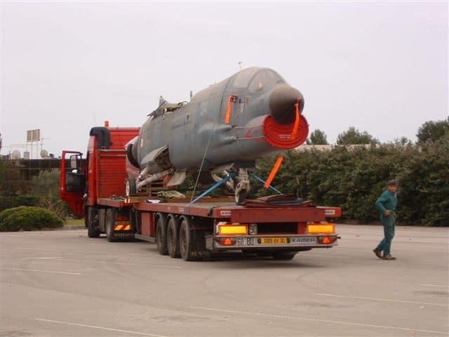 A large military fighter jet, appearing to be a Mirage 2000, is strapped onto the flatbed of a red heavy-duty truck. The jet…