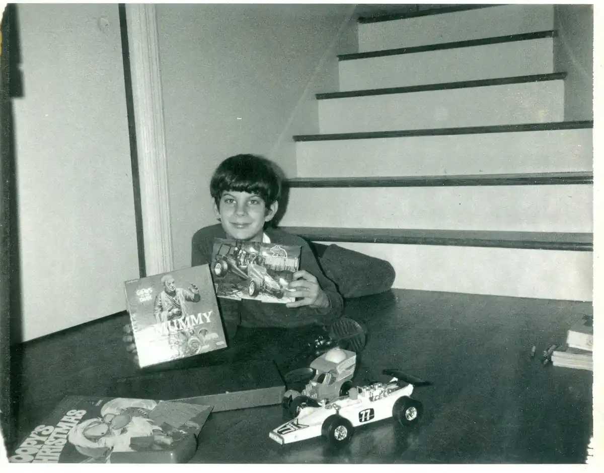 A black-and-white photo of a young boy sitting on a staircase landing, holding two toy packages labeled 'MUMMY' and 'Meyers…