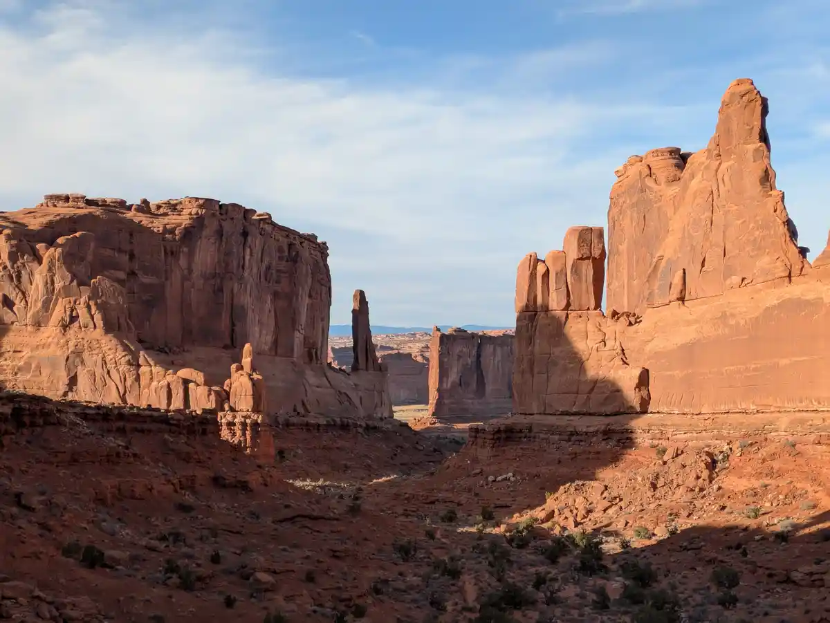 A scenic desert landscape featuring towering red rock formations under a partly cloudy blue sky, with sparse vegetation on…
