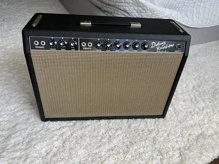 A vintage black Fender Deluxe Reverb guitar amplifier sits on a white shag carpet, its control panel showing knobs for…