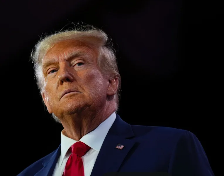 a close-up photo of Donald Trump looking upward with a serious expression, wearing a dark suit, white shirt, and red tie…