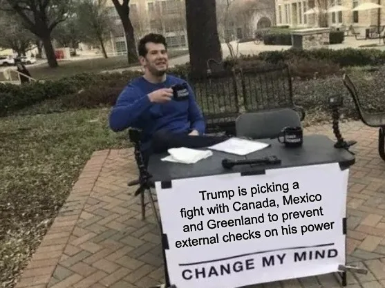 A man sits at a small outdoor table set up like a news desk, holding a mug, with a sign in front of him that reads 'Trump is…