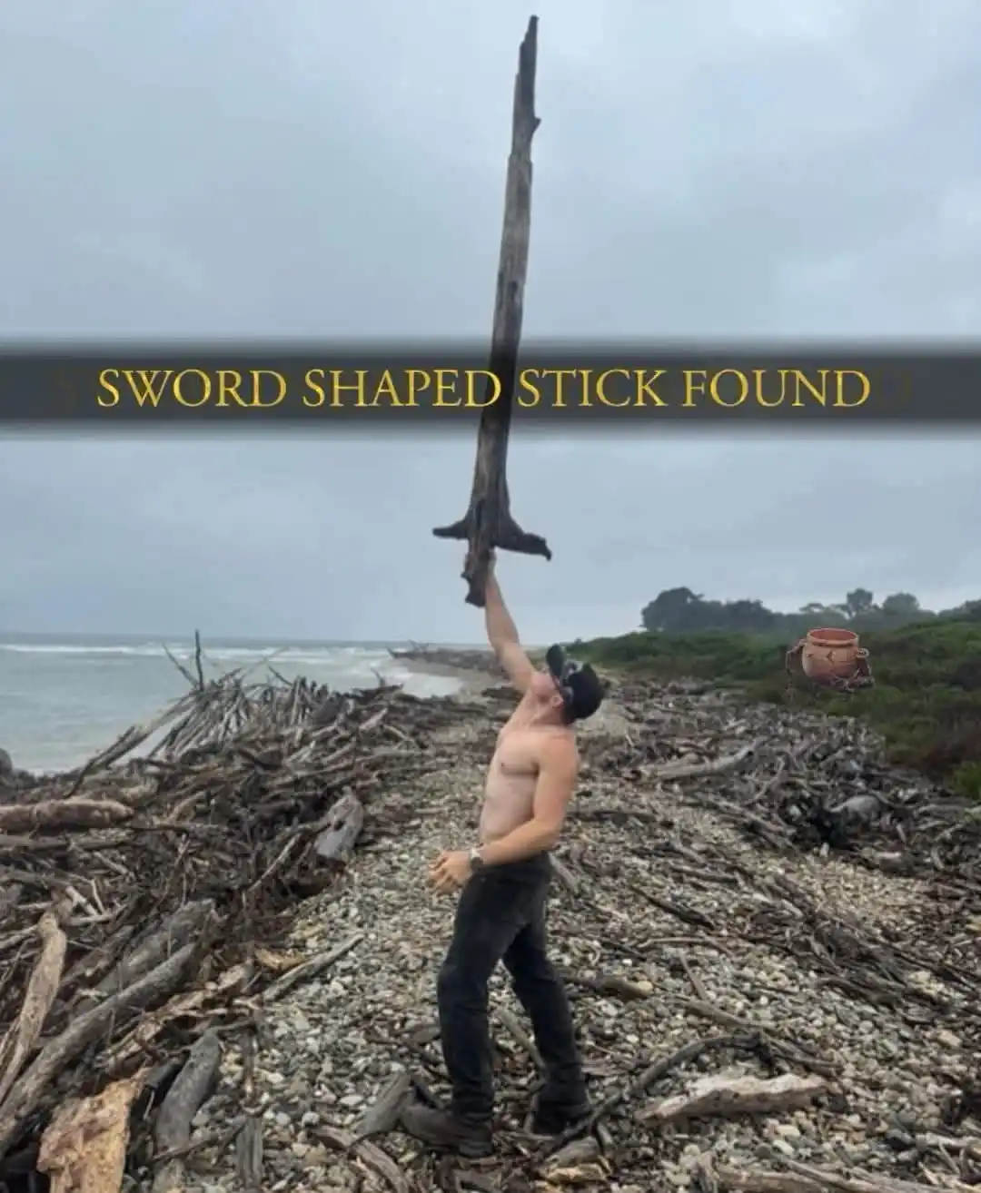 A shirtless man stands on a pebbly beach covered in driftwood, holding up a long, weathered piece of driftwood that vaguely…