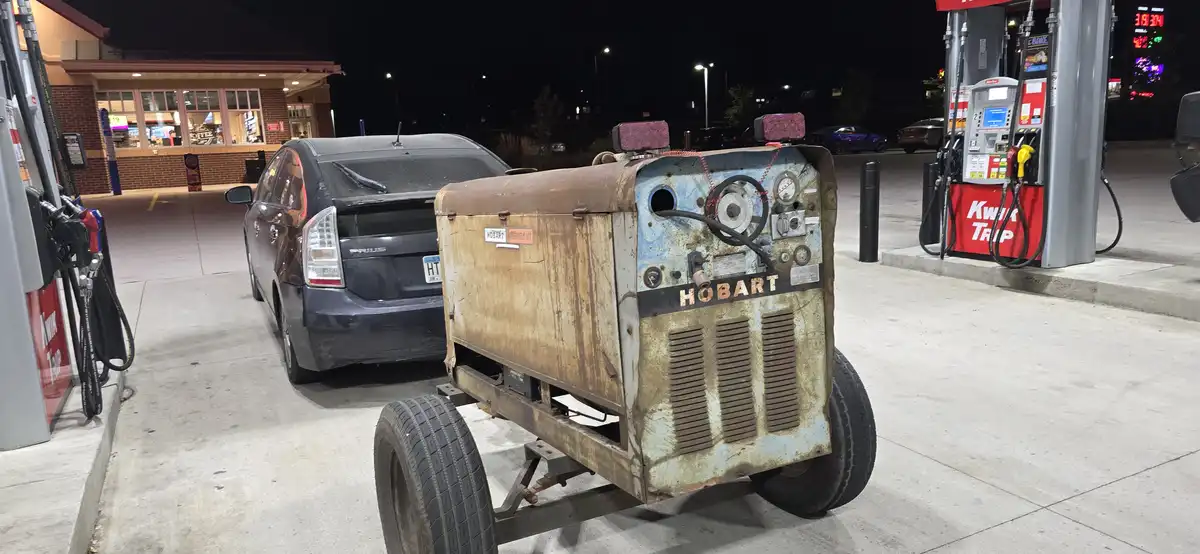 A rusty, wheeled Hobart welder machine is parked at a gas station pump next to a dark blue Toyota Prius at night. The welder…