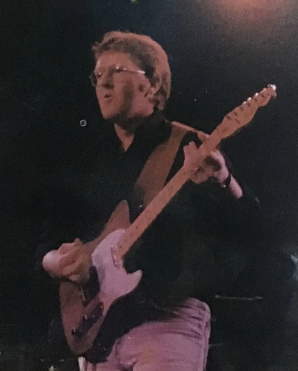 A man with glasses and short hair is playing a light-colored electric guitar on a dark stage, wearing a black shirt and…