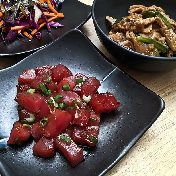 A close-up photo shows three black dishes on a wooden table: a central plate of raw, cubed red tuna topped with green…