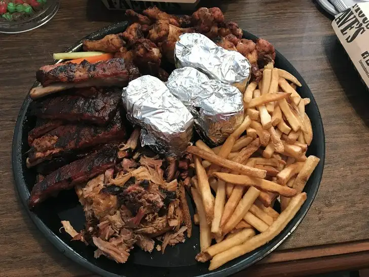 a large black plate piled high with various barbecue items including ribs, pulled pork, fried chicken wings, french fries…