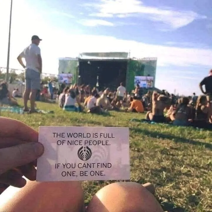 A person holds up a small white card with black text at a crowded outdoor concert or festival. The card reads 'THE WORLD IS…