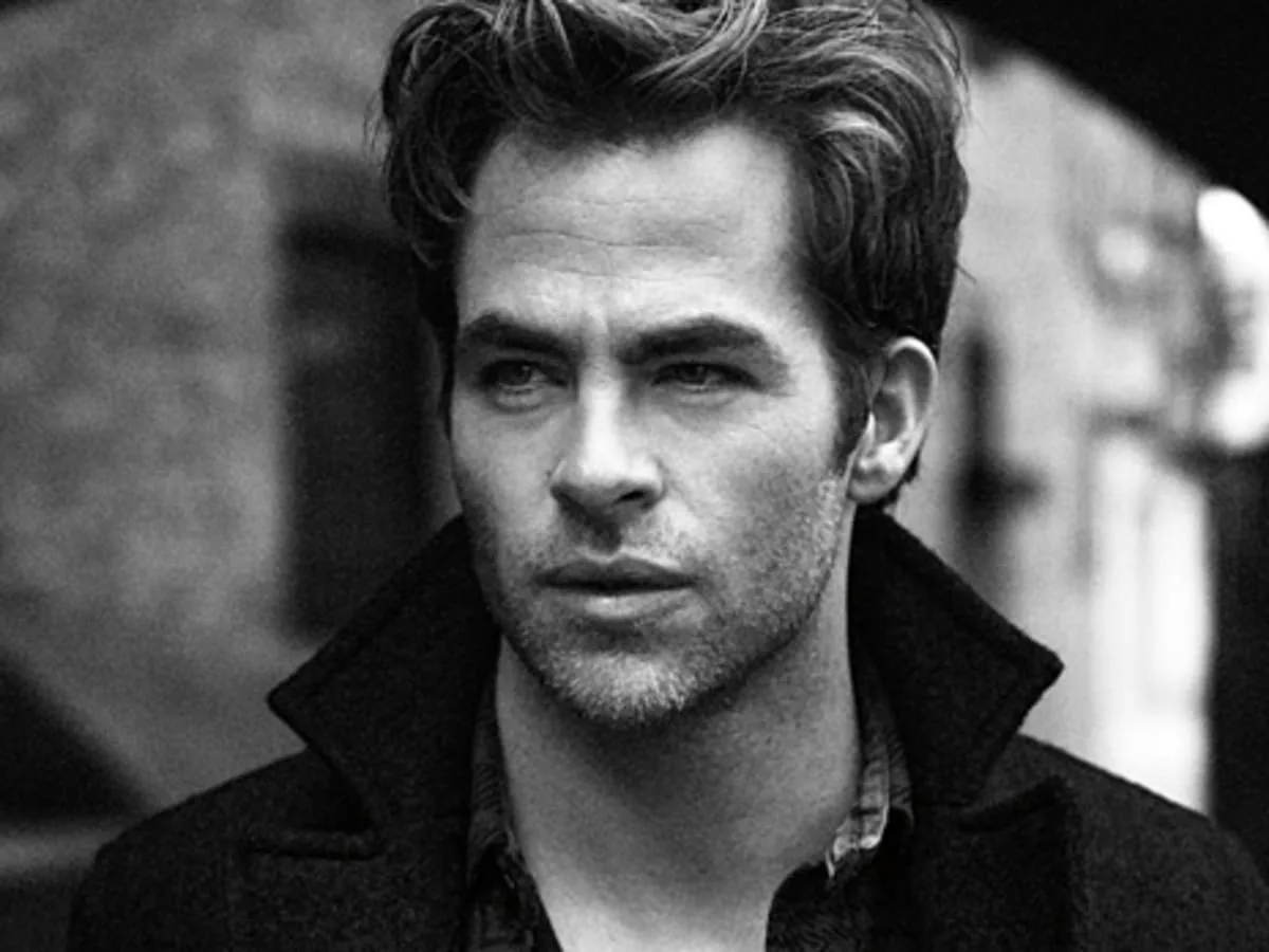 A black-and-white portrait of a man with styled hair and stubble, wearing a dark coat over a collared shirt, looking…