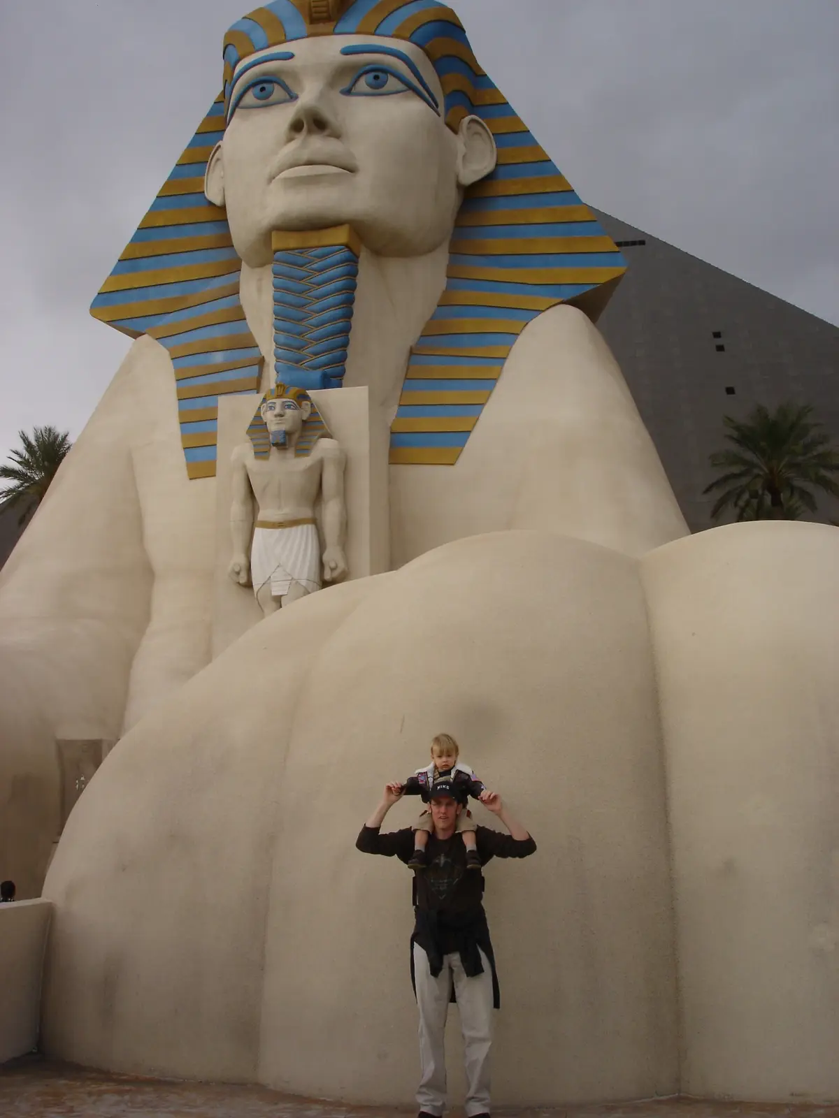 A man with a child on his shoulders stands in front of a giant, stylized Egyptian pharaoh statue with blue and gold striped…