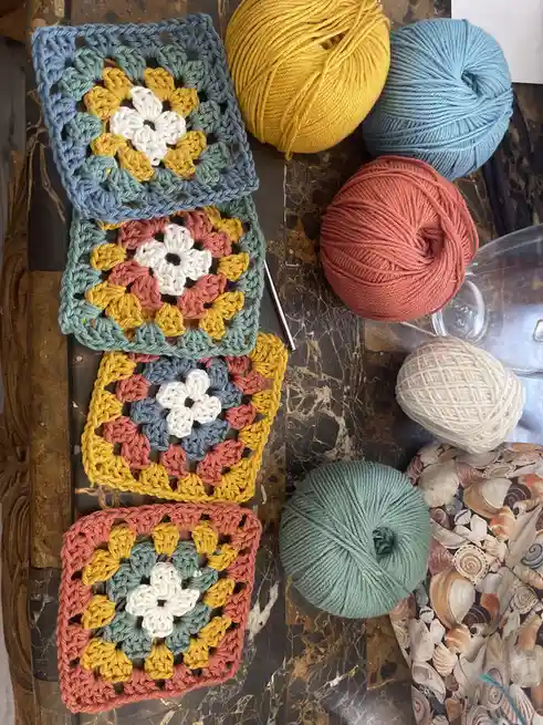 An overhead photo shows four colorful crocheted granny squares arranged vertically on a dark marble surface, surrounded by…