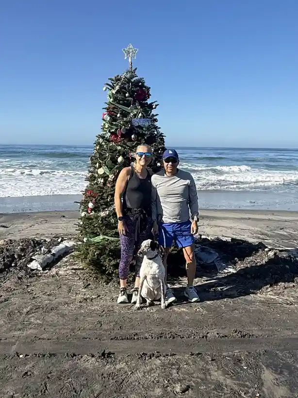 A man and woman wearing sunglasses and athletic clothing stand beside a decorated Christmas tree on a dark sandy beach with…