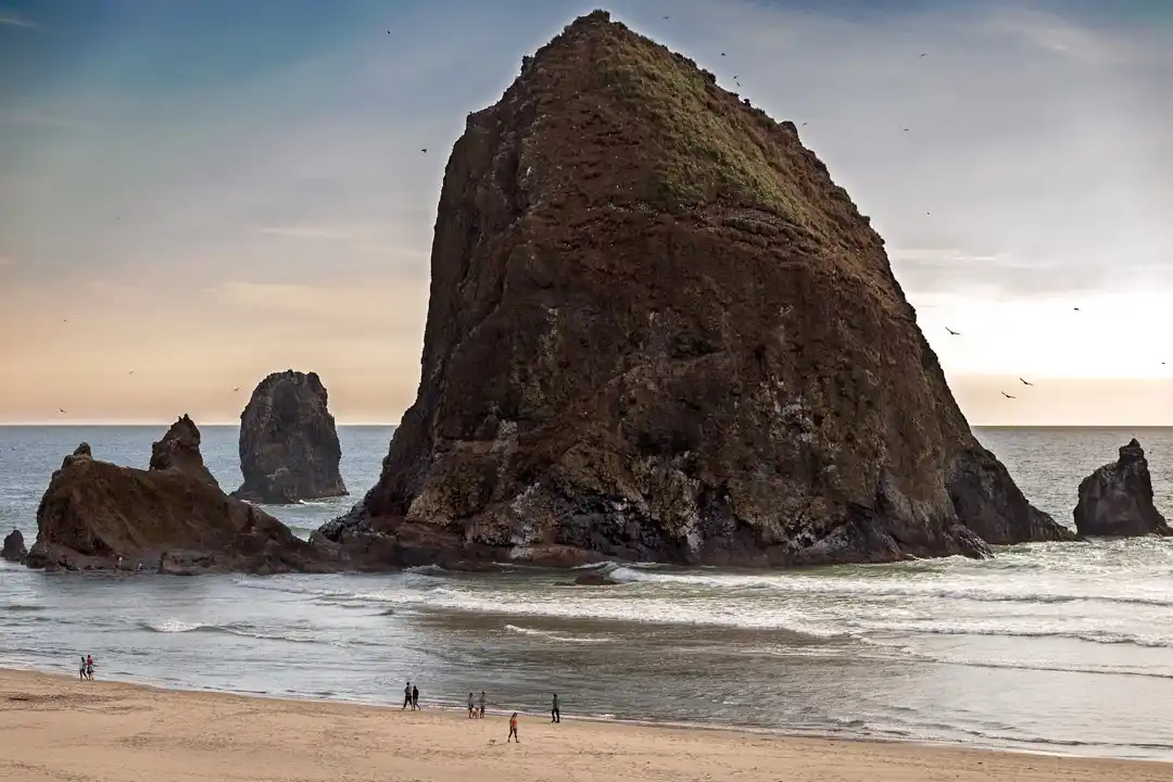 A scenic photo of a large, prominent sea stack on a beach with smaller rocks nearby and people walking on the shore. The…
