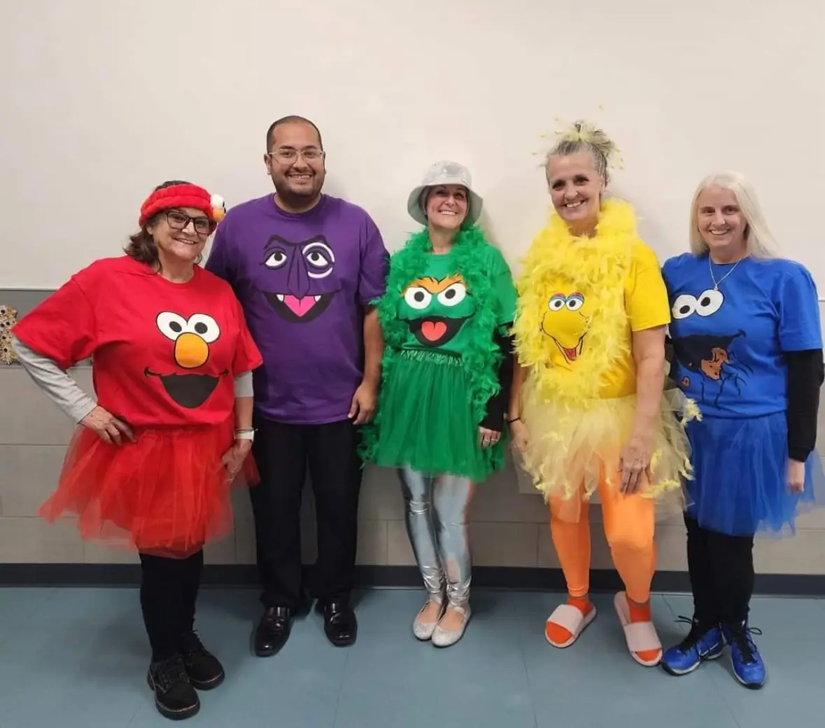 Five people pose together in costumes resembling Sesame Street characters: Elmo (red shirt, tutu), a purple-faced character…