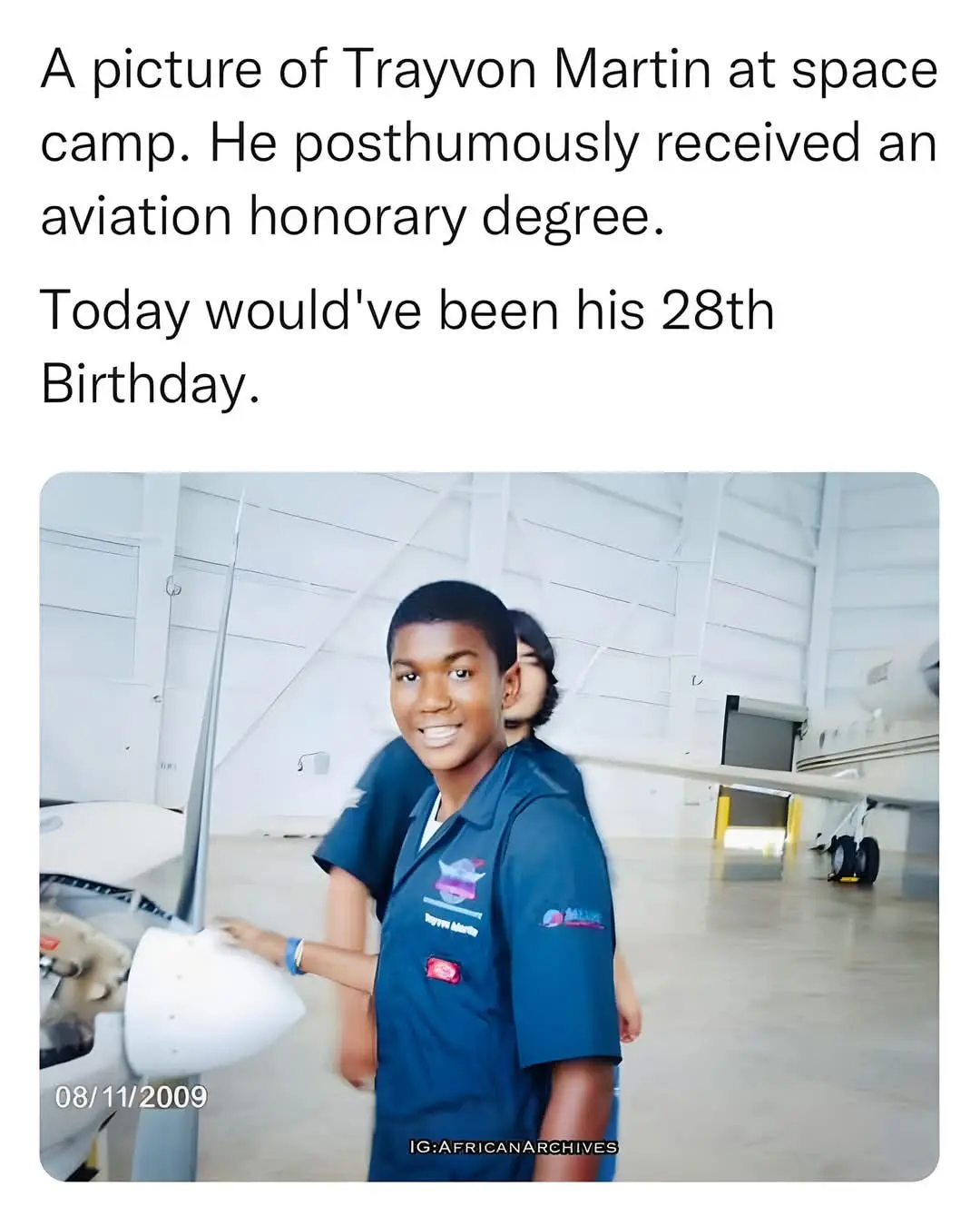 A young Black boy in a blue space camp uniform smiles at the camera while standing next to an airplane propeller in a…