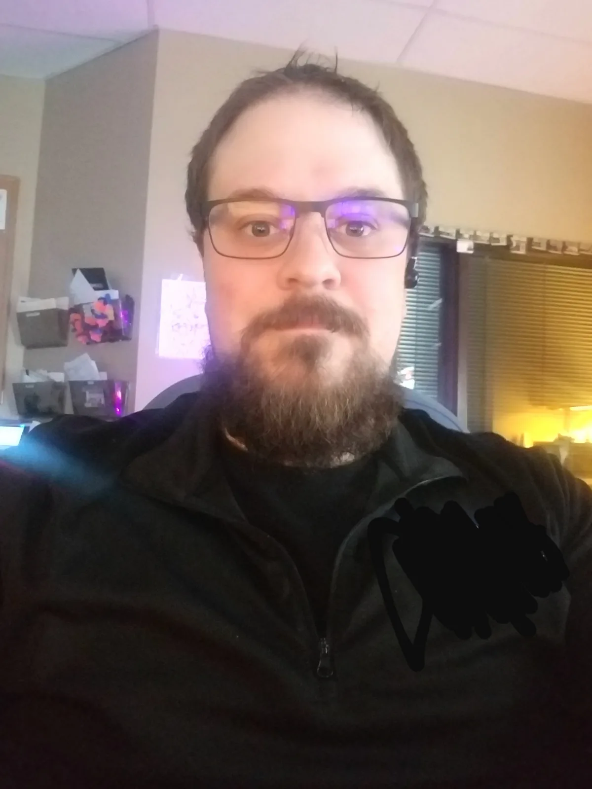 A man with glasses and a beard stares directly at the camera with a neutral expression, wearing a black zip-up jacket. A…