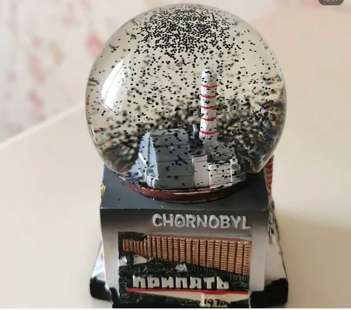 A snow globe with a miniature Chernobyl nuclear power plant inside, filled with black specks resembling radioactive dust or…
