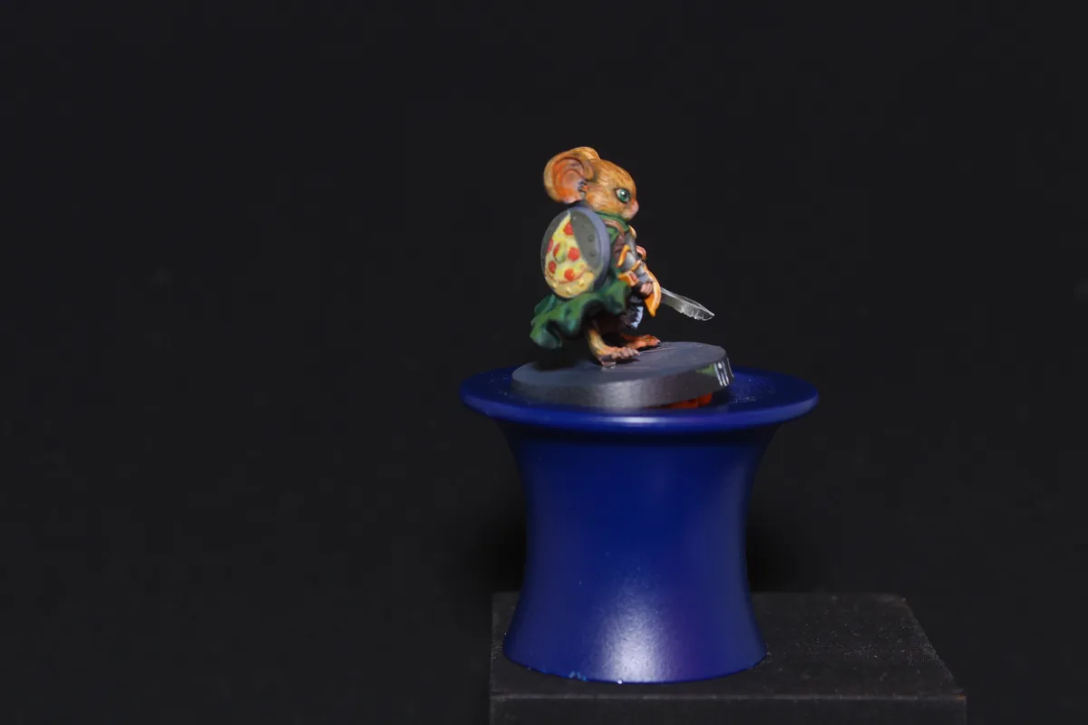 A small, painted mouse figurine dressed as a warrior with a shield and sword stands atop a blue top hat against a black…