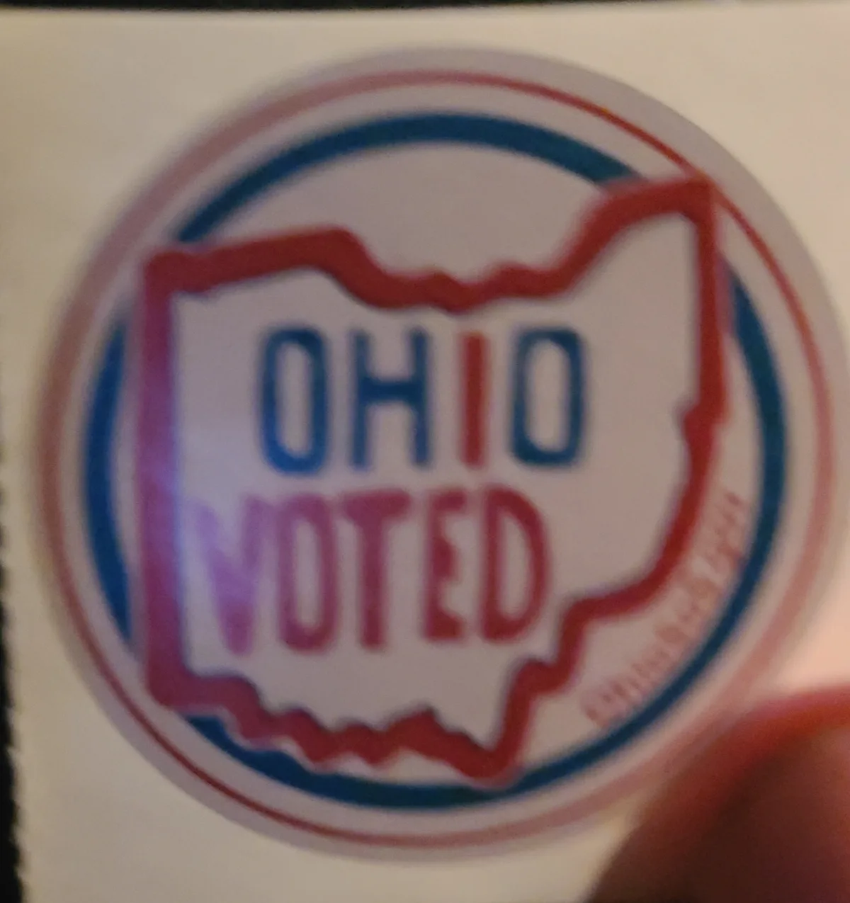 A blurry close-up of a circular sticker showing the outline of the state of Ohio in red, with the words 'OHIO' in blue and…