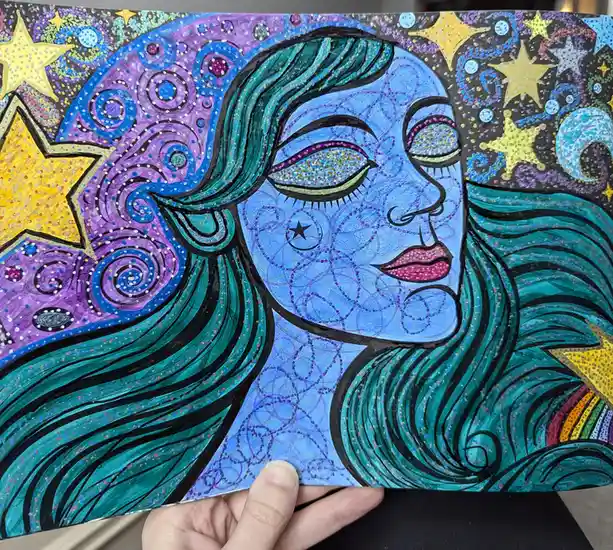 A hand holds up a colorful, glittery drawing of a woman's face in profile, her skin blue, hair teal, and eyes sparkling with…