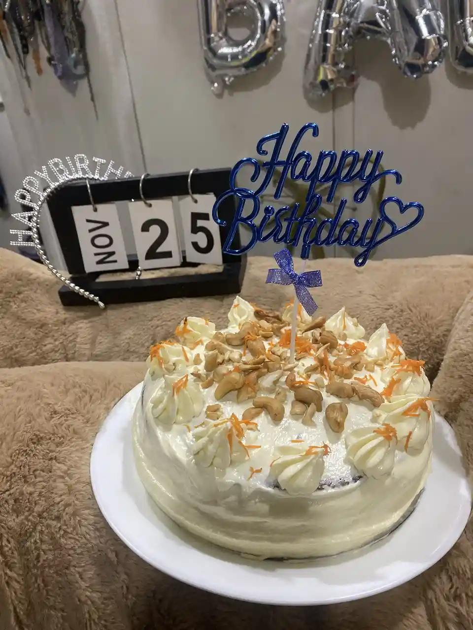 A birthday cake with white frosting, cashews, and orange zest sits on a white plate atop a brown fluffy surface. Behind it…