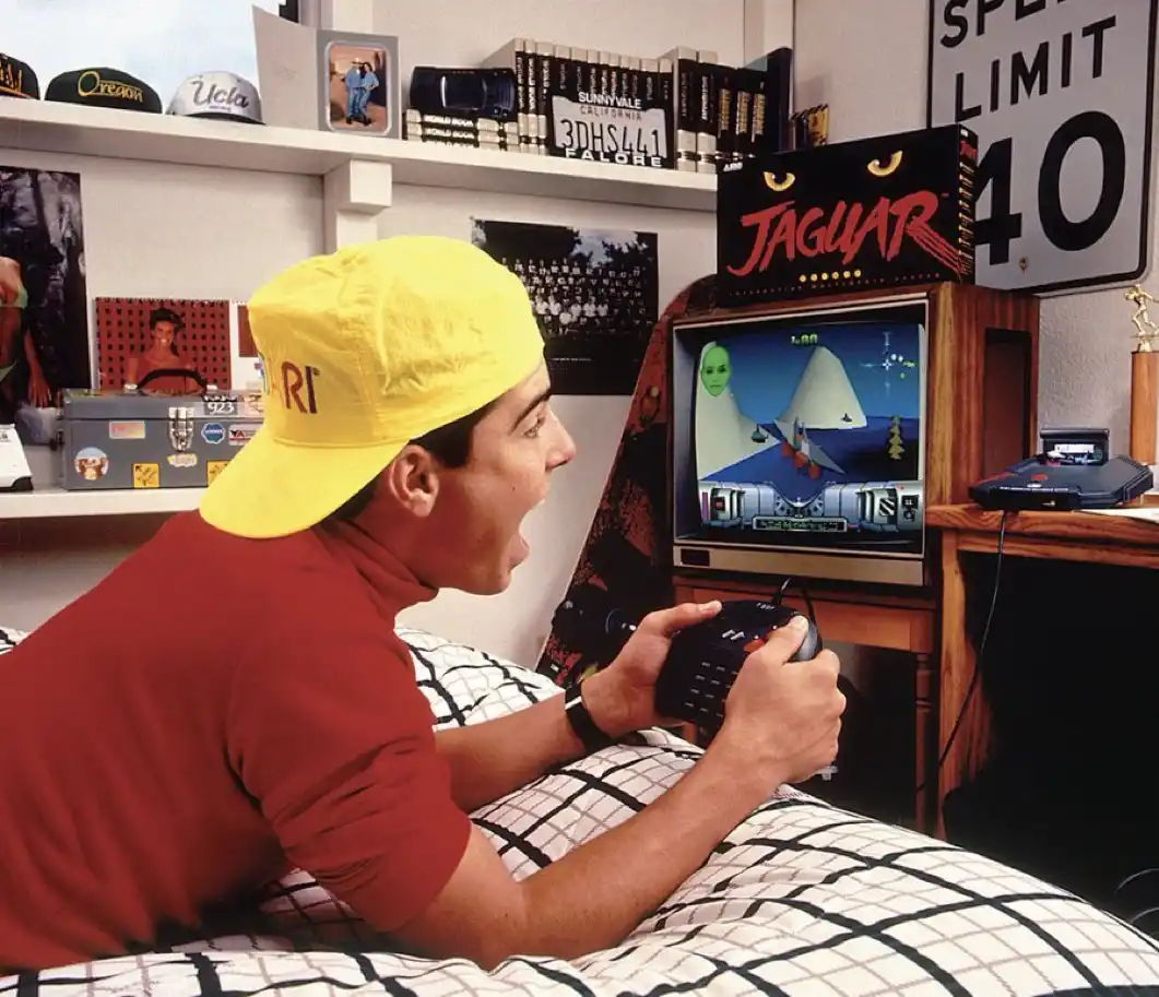 A young man in a yellow Atari cap and red shirt lies on a checkered bed, intensely playing a video game on a vintage Atari…