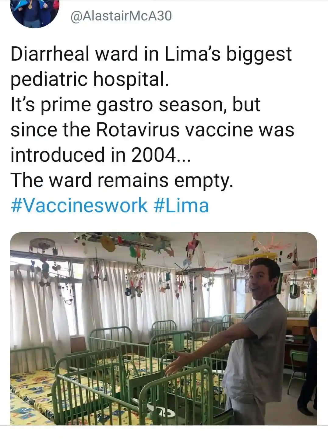 A man in medical scrubs stands in an empty pediatric hospital ward filled with green cribs and colorful mobiles, gesturing…