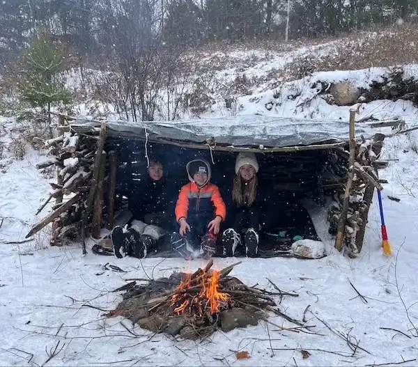 Three children sit inside a rustic, snow-covered lean-to shelter in a snowy forest, smiling at the camera. In front of them…