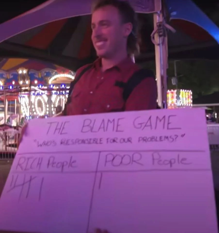 A man at a nighttime carnival holds a handwritten sign titled 'THE BLAME GAME' with the question 'WHO'S RESPONSIBLE FOR OUR…