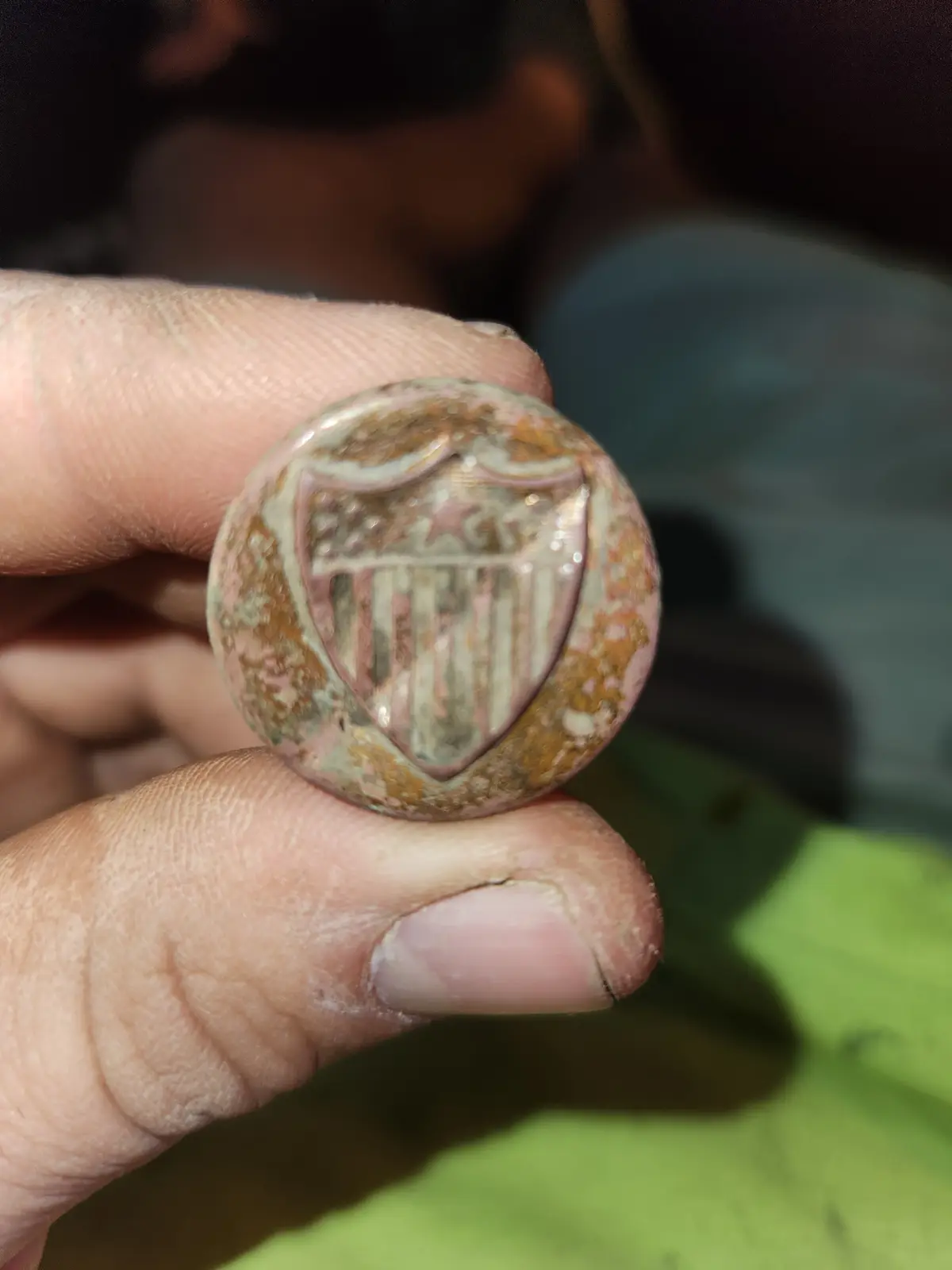 A close-up photo shows a hand holding a small, round, heavily corroded metal object with a faded American flag shield…